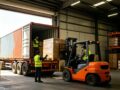 Overseas warehouse loading dock, workers and forklift unloading pallet cartons from container truck, inbound transshipment for cross-border cargo