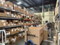 Real‑life photo of US overseas warehouse, warehouse staff sorting parcels, full storage shelves filled with cartons, DHL eCommerce large box, cross‑border seller inventory fulfillment scene
