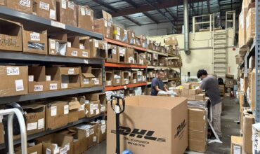 Real‑life photo of US overseas warehouse, warehouse staff sorting parcels, full storage shelves filled with cartons, DHL eCommerce large box, cross‑border seller inventory fulfillment scene