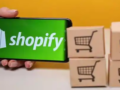 Hand holding a mobile phone with Shopify logo next to cross-border shipping boxes
